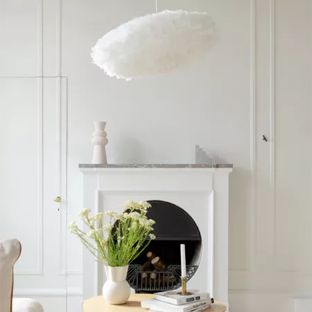 Darc — Une cheminée blanche en marbre avec un foyer noir, surmontée d’un lustre en forme de nuage blanc. À gauche, un vase avec des fleurs séchées et