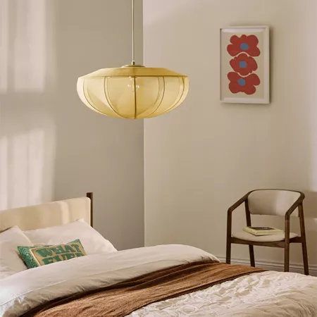 Darc — Une chambre lumineuse avec une suspension en forme de cloche jaune suspendue au-dessus d’un lit simple, recouvert d’un plaid beige et d’un cous