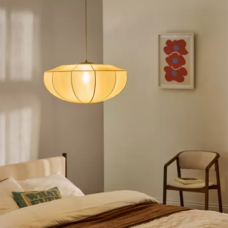 Darc — Une lampe suspendue en tissu beige clair, de forme ovale et légèrement bombée, éclaire une chambre minimaliste avec un lit fait et une couvertu