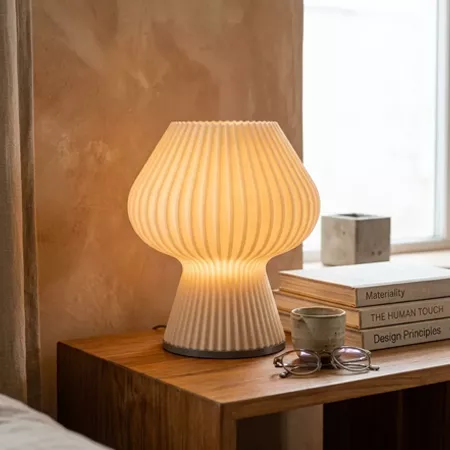 Darc — Une lampe design en forme de champignon blanc aux plis fins, posée sur une table en bois clair près d'une fenêtre. À côté, des livres sur le de