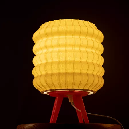 Darc — Lampe design en tissu jaune doré en forme de nid d'abeille, posée sur un pied rouge triangulaire, émettant une lumière chaude sur fond noir.