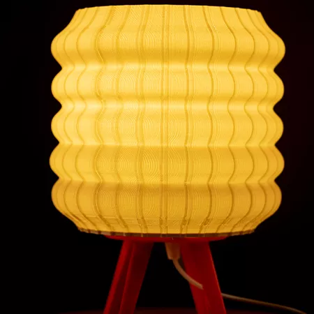 Darc — Lampe design composée d’un abat-jour en tissu jaune plissé et strié, posée sur un pied rouge en métal triangulaire, émettant une lumière douce