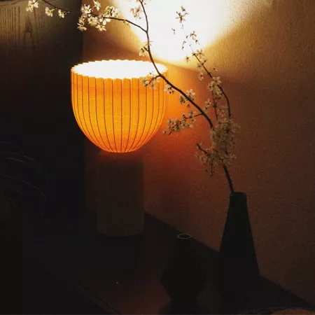 Darc — Une lampe design en forme de panier orange éclairée, posée près d'une branche fleurie blanche aux fleurs délicates, créant une ambiance chaleur