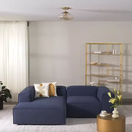 Darc — Un salon lumineux avec un canapé bleu profond en forme de L, des coussins blancs et dorés, une table basse ronde en bois clair, une étagère dor